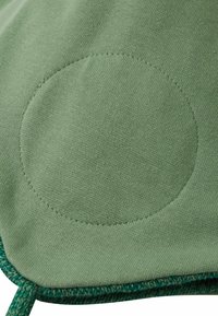 Zielona tkanina z okrągłą łatą i widocznym przeszyciem. Tekstura jest gładka, z lekko podniesionym brzegiem w ciemniejszym odcieniu zieleni.