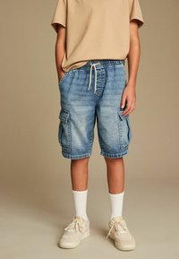 Pantalones cortos cargo de mezclilla con cintura elástica y cordón, con dos bolsillos laterales y un lavado azul claro. Combinados con calcetines blancos y zapatillas deportivas.