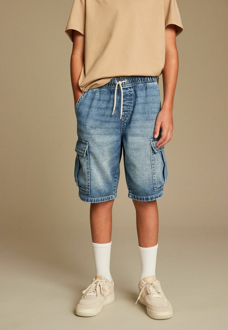 Pantalones cortos cargo de mezclilla con cintura elástica y cordón, con dos bolsillos laterales y un lavado azul claro. Combinados con calcetines blancos y zapatillas deportivas.
