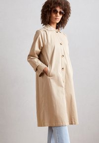 Geacă de trench de culoare crem, cu o croială lejeră, guler mare și închidere cu buton, având buzunare laterale și mâneci lungi, combinată cu blugi.