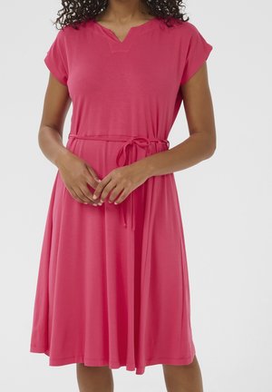 Femme portant une robe rose courte jusqu'aux genoux, manches courtes, col en V et ceinture nouée à la taille sur un fond uni.