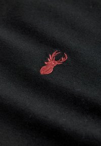 Schwarzer Stoff mit glatter Textur, der ein kleines, rotes gesticktes Hirschdesign zeigt, das filigrane Geweihe präsentiert.