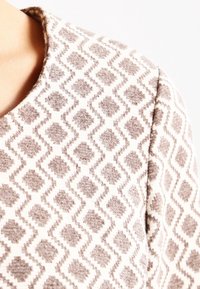 Pull en peluche de couleur claire avec un motif texturé présentant des formes de losanges brunes. Encolure ronde et matériau en tricot doux.