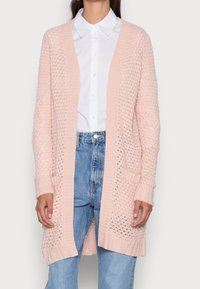 Mujer vestida con un cárdigan de punto abierto en rosa claro sobre una camisa blanca con cuello y jeans de tiro alto azules, de pie contra un fondo liso.