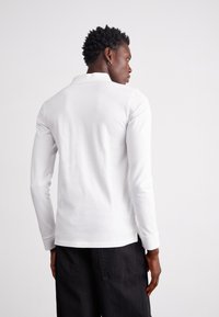 Gaišais kokvilnas polo krekls ar garām piedurknēm un apkārtni, veidots pieguļoši, ar gludu tekstūru. Valkāts ar tumšiem biksēm.