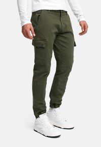 Homme portant un pantalon cargo vert olive avec des poignets élastiques et des baskets montantes blanches, debout devant un fond blanc.
