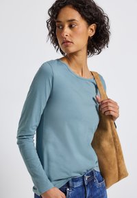 Žena s kudrnatými vlasy, oblečená ve světle modré košili s dlouhým rukávem a modrých džínách, drží hnědou semišovou tašku přes pravé rameno.