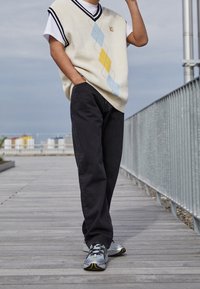 Osoba stojąca na drewnianym deptaku, ubrana w biały sweter w romby, czarne spodnie i szare sneakersy, z ręką w kieszeni.