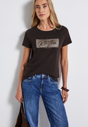 Woman wearing black graphic t-shirt with "The Rhythm of Nature," blue jeans, brown bracelets, and holding a brown handbag against white wall.