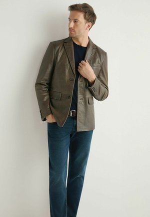 Mężczyzna ubrany w ciemnozieloną skórzaną marynarkę na czarnej koszuli i niebieskich dżinsach, stojący przy białej ścianie, patrzący na bok z jedną ręką na marynarce.