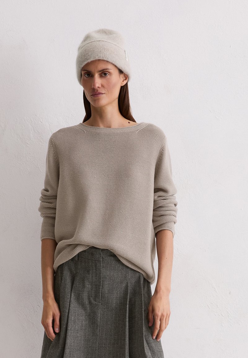 Beige gebreide trui met een brede halslijn, volle mouwen en een oversized fit, gecombineerd met een grijze geplooide rok. Zachte textuur, casual stijl.