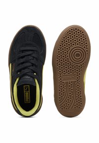 Zwarte suède sneakers met gele accenten, voorzien van een ronde neus en een getextureerde bruine rubberen zool met cirkelvormige patronen en branding.