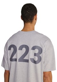 Mand med kort krøllet hår iført en lysegrå t-shirt med store mørkegrå tal "223" på ryggen.