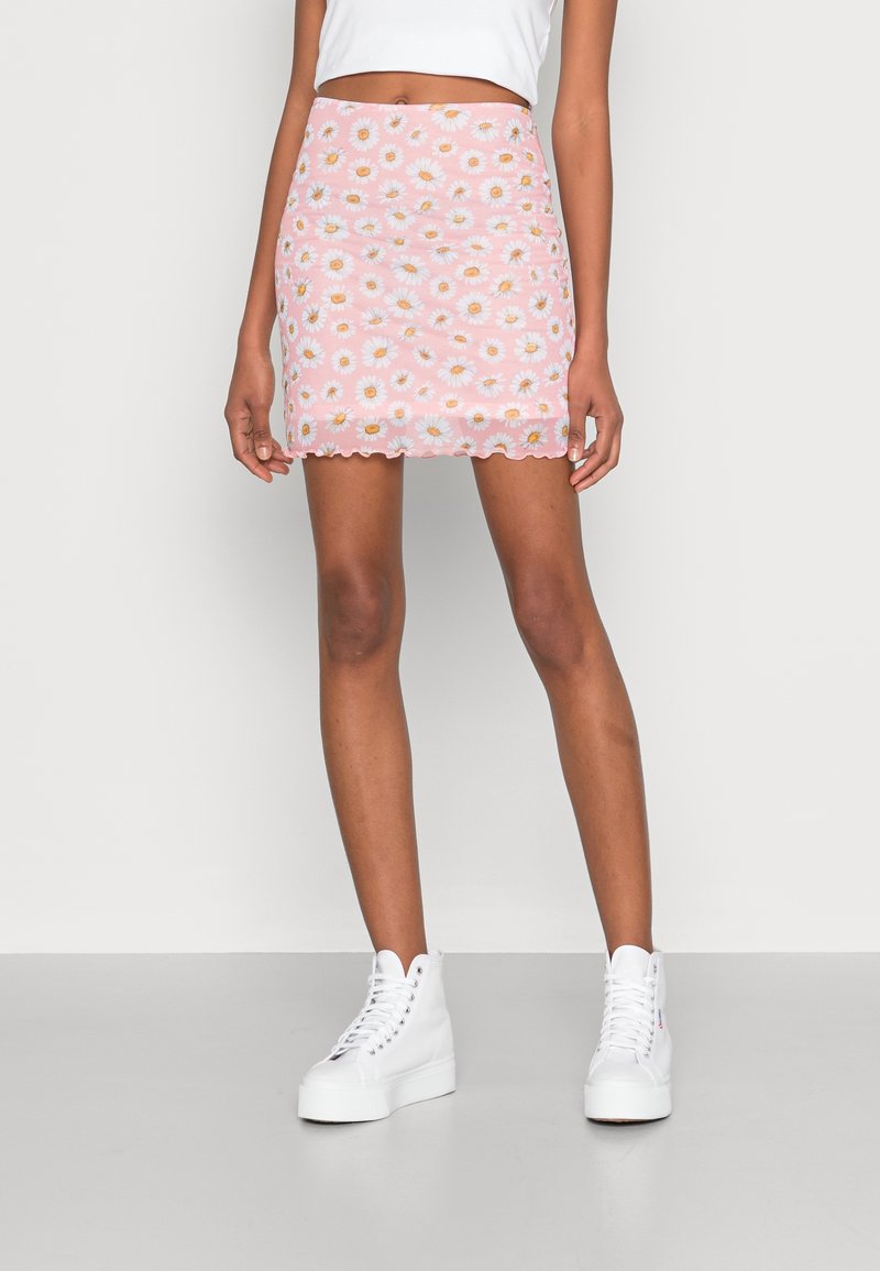 Mini-jupe rose avec des motifs de marguerites blanches, ourlet festonné, coupe ajustée, associée à des baskets montantes blanches et un top court blanc.