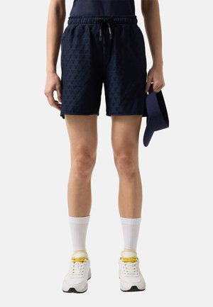 Personne portant un short bleu marine texturé avec cordon de serrage, des chaussettes blanches, des baskets blanches avec des lacets jaunes, tenant une visière bleu marine assortie.