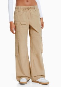Beige cargobroek met een losse pasvorm, een koordsluiting in de taille, meerdere zakken en een gladde katoenen textuur, gestyled met witte sneakers.