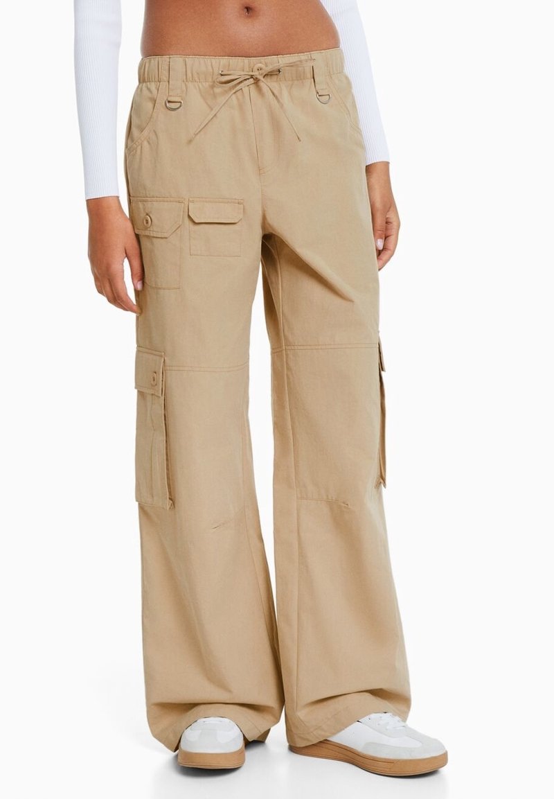 Beige cargobroek met een losse pasvorm, een koordsluiting in de taille, meerdere zakken en een gladde katoenen textuur, gestyled met witte sneakers.