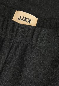 Fascia elastica nera con una finitura testurizzata e luccicante. L'etichetta riporta "JJXX". Il materiale è morbido, con un leggero motivo a coste e lucentezza.