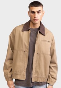 Tan zip-up jacket with a brown corduroy collar, two front pockets, elastic cuffs, and a relaxed fit worn over a gray shirt.