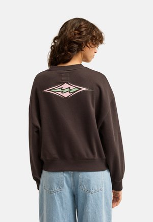 Personne aux cheveux bouclés portant un sweat-shirt marron foncé avec un motif géométrique rose et vert dans le dos et un jean bleu clair.