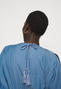 Vaate denim-paita vaaleansinisena, jossa on rypytetty selkä ja punottu nauha sinivalkoisilla tupsuilla. Pehmeä rakenne ja rento malli.