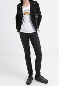 Černá semišová motorkářská bunda se stříbrnými detaily na bílem tričku s grafikou pizzy. Černé slim fit kalhoty a černé boty.