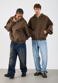 Rjava zip-up kapuca, sproščenega kroja, prevelike rokave. V kombinaciji z modrimi jeans hlačami; eden svetlejši, drugi temnejši z izbledelim detajlom. Črne čevlje z dodatki.