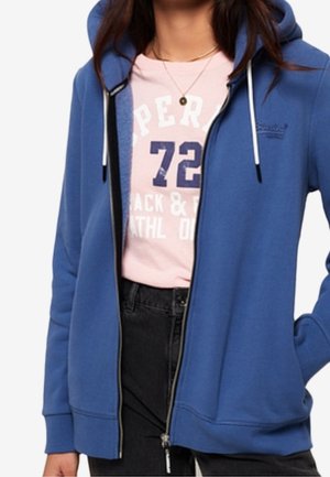 Niebieska bluza z kapturem na suwak z białymi sznurkami, noszona na jasnoróżowym graficznym t-shircie i czarnych spodniach.