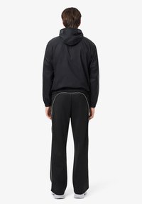 Zwarte windbreaker met capuchon, elastische manchetten en zoom; gecombineerd met zwarte trainingsbroeken met witte zijstrepen en grijze sneakers.