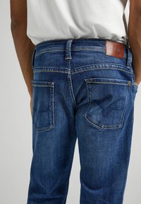 Jeans de mezclilla con un lavado azul oscuro, con bolsillos traseros, costuras en contraste y una etiqueta de cuero marrón en la cintura, textura suave.