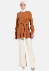 Haut marron à manches longues avec un détail noué, associé à un pantalon blanc évasé. Tissu lisse, coupe décontractée et design drapé subtil.