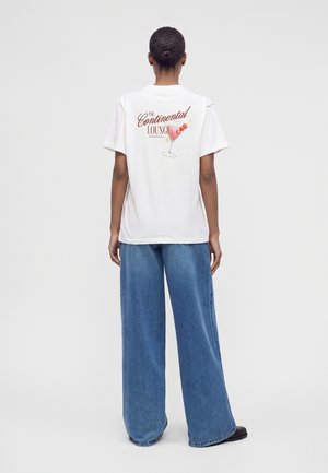 Personne avec les cheveux attachés en chignon porte un t-shirt blanc avec un verre à cocktail et le texte "The Continental Lounge" dans le dos et un jean bleu large.