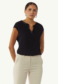 Zwarte mouwloze top met een lichte glans, met een ingehaakte kraag en een gladde textuur, gecombineerd met lichtbeige gepaste broeken.