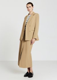 Blazer beige su misura con ampi risvolti e bottoni doppi, abbinato a una gonna coordinata con uno spacco frontale. Mocassini neri completano il look.