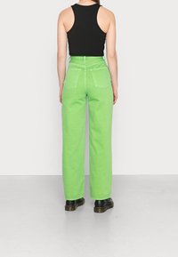 Vaqueros verdes de pierna ancha con cintura alta, dos bolsillos traseros y detalles de costura visibles, combinados con una blusa sin mangas negra y zapatos negros.