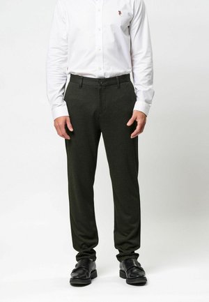Mężczyzna ubrany w ciemnozielone, dopasowane spodnie, czarne eleganckie buty oraz białą koszulę na długi rękaw z małym haftowanym logo.