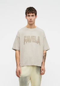 Tricou grafic bej cu cuvântul "FAVELA" scris cu litere îndrăznețe. Prezintă o croială lejeră și mâneci scurte, fiind fabricat dintr-un material texturat și moale.