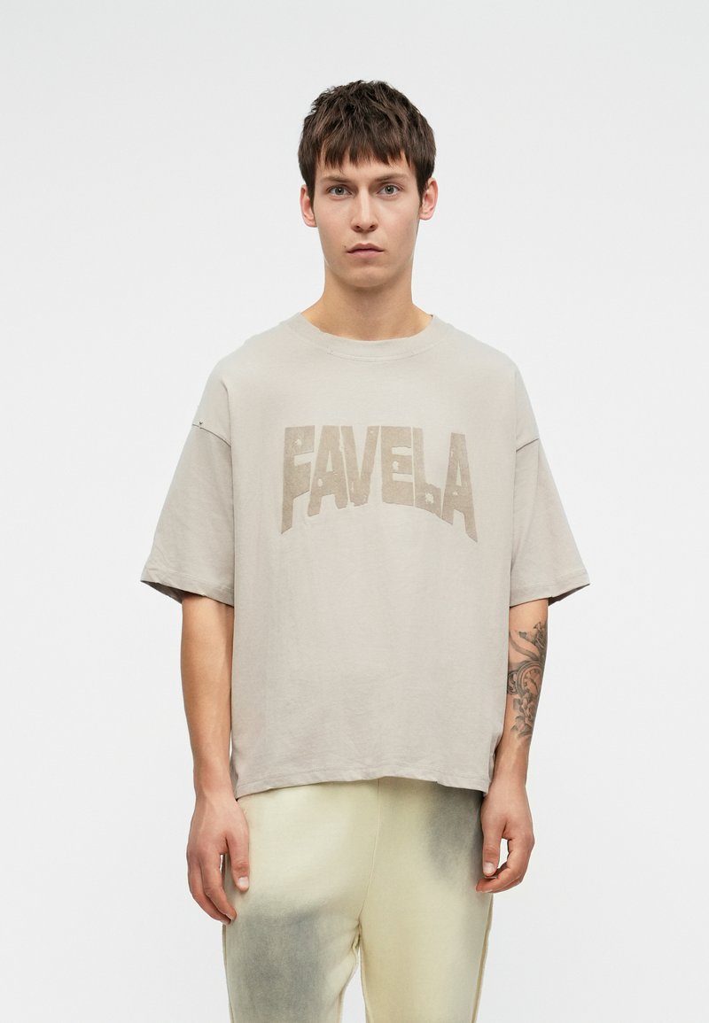 Tricou grafic bej cu cuvântul "FAVELA" scris cu litere îndrăznețe. Prezintă o croială lejeră și mâneci scurte, fiind fabricat dintr-un material texturat și moale.