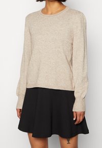Pull en maille beige avec un col rond et des poignets côtelés, associé à une jupe évasée noire. Texture douce, coupe décontractée et design simple.