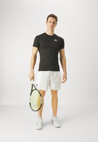 Camiseta atlética negra de manga corta con un logo blanco, combinada con pantalones cortos blancos y zapatillas de tenis grises, sosteniendo una raqueta de tenis.