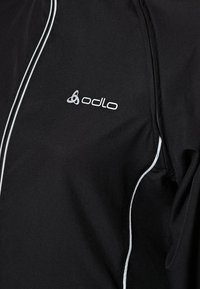Top deportivo negro con detalles reflectantes en blanco, cremallera completa y el logo de Odlo en la zona del pecho. Textura de tela suave.