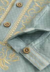 Chemise brodée en tissu texturé bleu clair, ornée de motifs en fil doré et de boutons en métal marron pour une touche décorative.