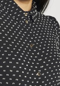 Blouse noire avec un motif texturé de points blancs, dotée d'un col avec revers et de détails de boutons dorés sur le devant.