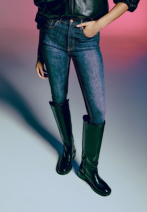 Persona con vaqueros ajustados azules, botas de cuero negras hasta la rodilla y una chaqueta de cuero negra sobre un fondo degradado rosa y gris.