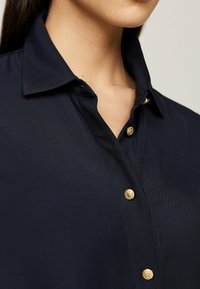 Camicia blu navy con colletto strutturato, presenta bottoni dorati e una finitura in tessuto strutturato, che mette in evidenza motivi a righe diagonali.