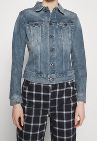 Denim jacket in light blue, featuring a fitted shape, classic collar, button closure, and two chest pockets with black buttons.