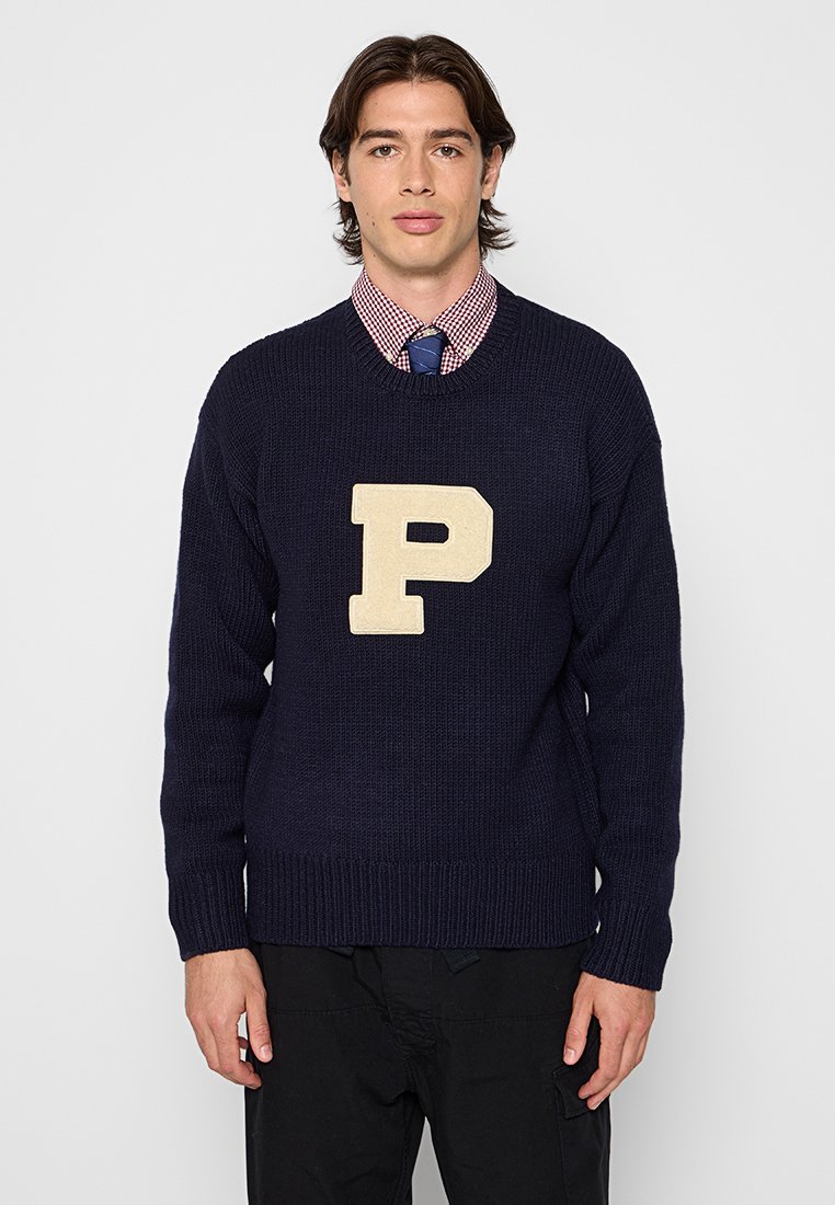 Marineblauer Strickpulli mit einem cremefarbenen "P"-Logo auf der Vorderseite. Hat einen runden Ausschnitt und gerippte Säume, getragen über einem karierten Hemd.