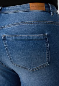 Blue denim jeans with a smooth texture, featuring a brown leather label reading "ONLY CARMAKOMA" and a standard back pocket design.