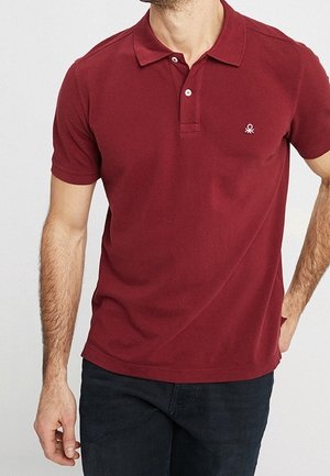 Mand iført en kortærmet, burgunder polo med krave og to knapper, kombineret med mørke bukser mod en ensfarvet baggrund.