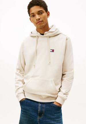 Felpa beige realizzata in tessuto morbido, con tasca frontale e cappuccio con lacci. Include una piccola patch logo rossa e blu sul petto.